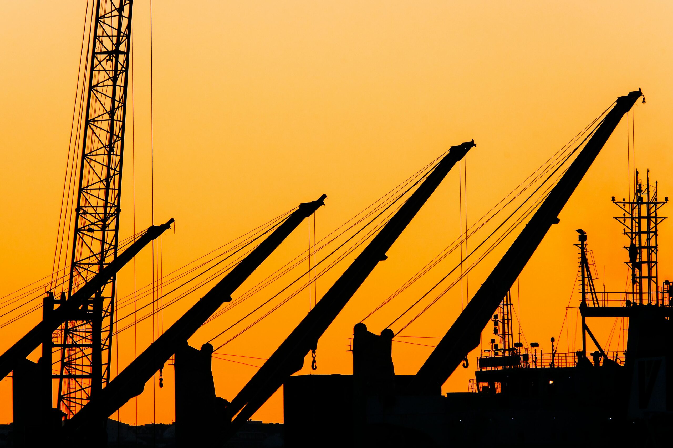 Silhouetted cranes and masts create a striking industrial skyline against a vibrant sunset backdrop.